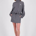 A woman modeling a black and white gingham mini jacket with a high collar, long sleeves, and a drawstring waist, paired with black flip-flop sandals against a light gray studio background.
