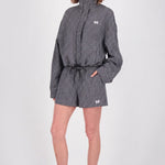 A model stands against a light gray studio background wearing a black and white gingham patterned high-neck jacket and matching shorts with a drawstring waist, paired with black flip-flops.