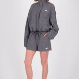 A model stands against a light gray studio background wearing a black and white gingham patterned high-neck jacket and matching shorts with a drawstring waist, paired with black flip-flops.