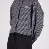 A model wearing a black and white gingham cropped jacket with a high collar and a small embroidered logo on the chest, paired with black trousers, against a light gray background.
