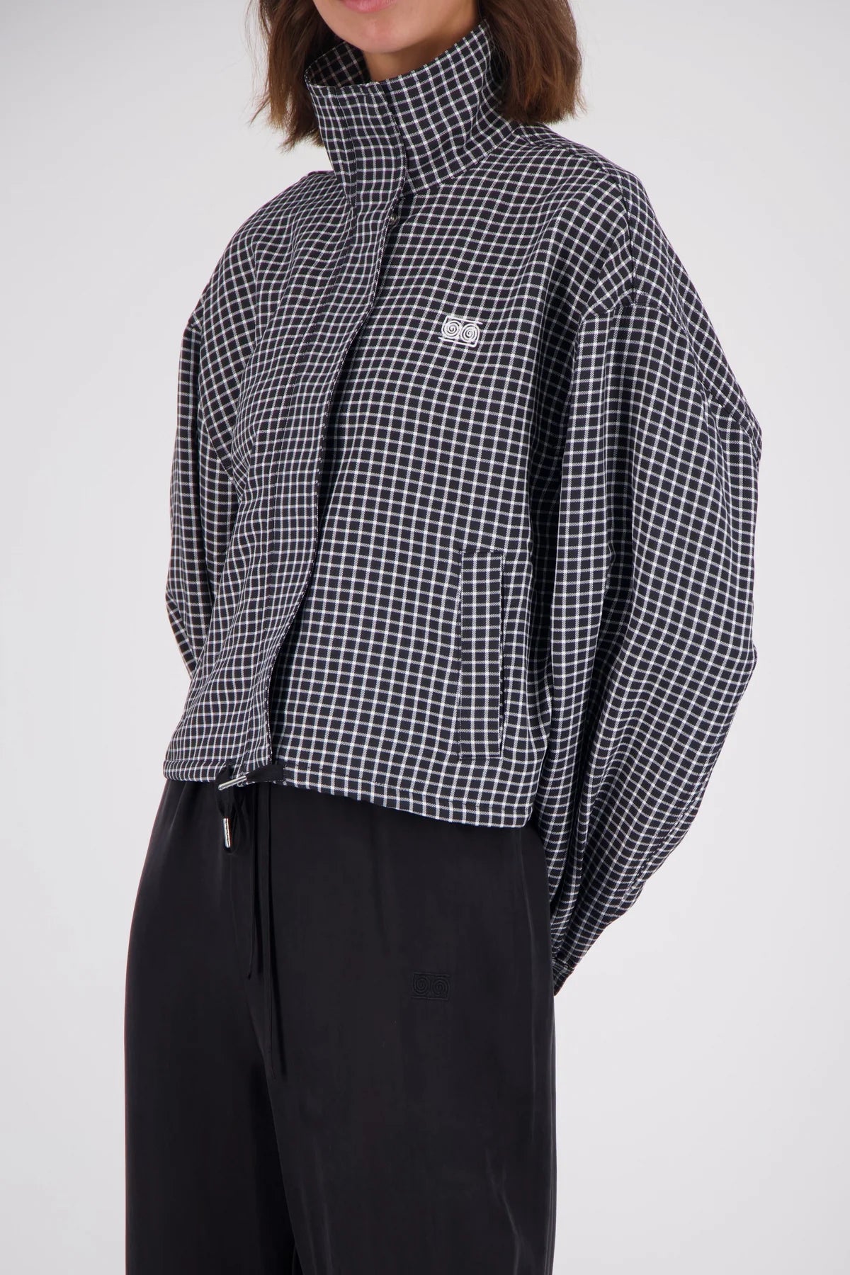 A model wearing a black and white gingham cropped jacket with a high collar and a small embroidered logo on the chest, paired with black trousers, against a light gray background.