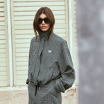 A woman wearing a black and white houndstooth mini jacket and matching shorts with a black tie belt, styled with black sunglasses, standing in front of a beige shuttered wall.