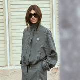 A woman wearing a black and white houndstooth mini jacket and matching shorts with a black tie belt, styled with black sunglasses, standing in front of a beige shuttered wall.