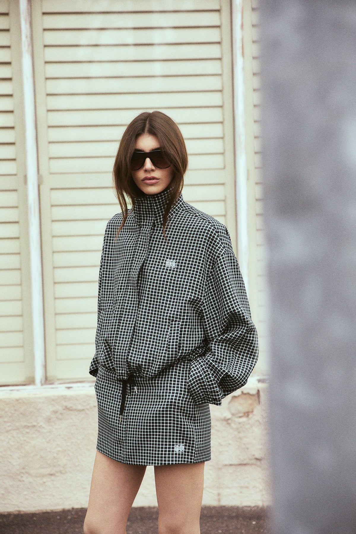 A woman wearing a black and white houndstooth mini jacket and matching shorts with a black tie belt, styled with black sunglasses, standing in front of a beige shuttered wall.