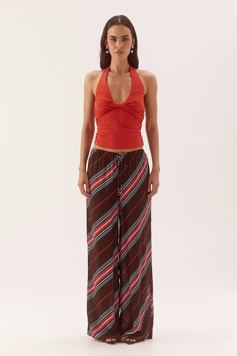 A woman modeling a sleeveless halter top in rust red paired with wide-leg pants featuring diagonal stripes in brown, red, and light blue, against a plain light gray studio background.