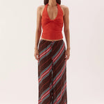 A woman modeling a sleeveless halter top in rust red paired with wide-leg pants featuring diagonal stripes in brown, red, and light blue, against a plain light gray studio background.