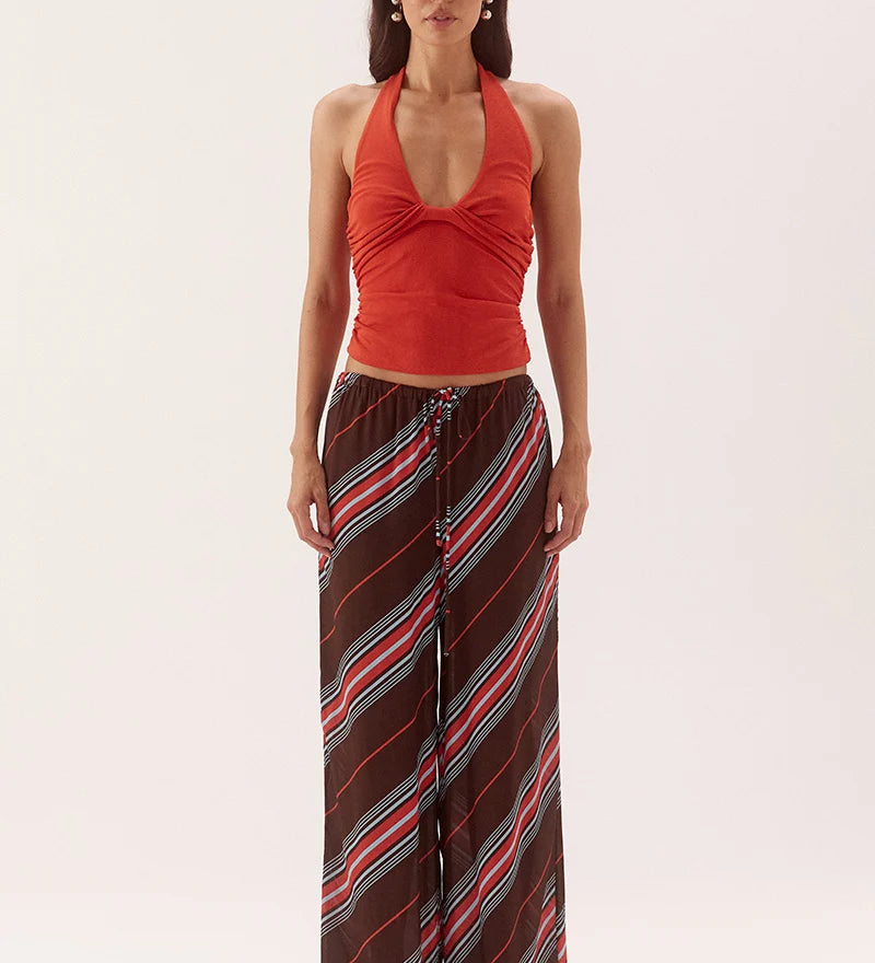 A woman modeling a sleeveless halter top in rust red paired with wide-leg pants featuring diagonal stripes in brown, red, and light blue, against a plain light gray studio background.