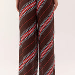 A person wearing loose, wide-leg pants with a diagonal striped pattern in dark brown, red, white, and gray, shown from the waist down against a plain light background.