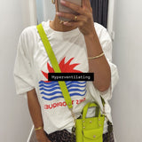 Person wearing a white t-shirt with a colorful graphic design, holding a neon green bag, and taking a mirror selfie.