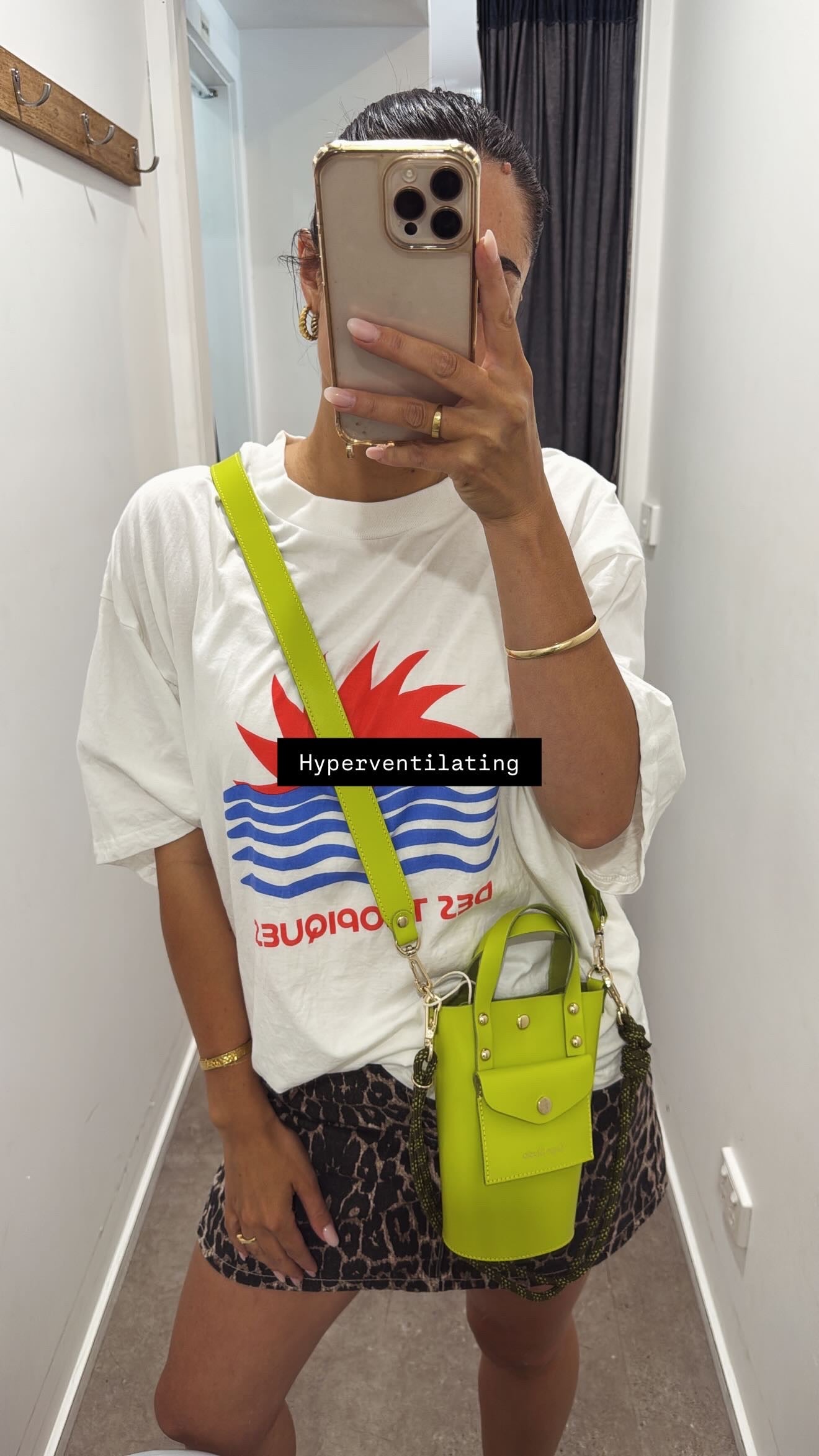 Person wearing a white t-shirt with a colorful graphic design, holding a neon green bag, and taking a mirror selfie.