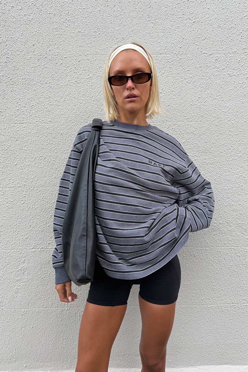 A woman wearing a gray and black horizontally striped long-sleeve sweatshirt with 'RAEF' printed on the chest, paired with black bike shorts, posing against a white textured wall.