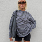 A woman wearing a gray and black horizontally striped long-sleeve sweatshirt with 'RAEF' printed on the chest, paired with black bike shorts, posing against a white textured wall.