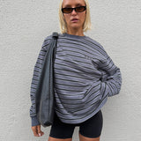 A woman wearing a gray and black horizontally striped long-sleeve sweatshirt with 'RAEF' printed on the chest, paired with black bike shorts, posing against a white textured wall.