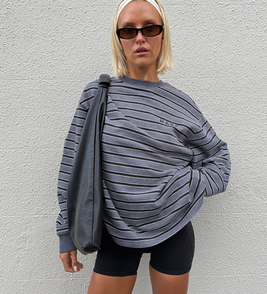 A woman wearing a gray and black horizontally striped long-sleeve sweatshirt with 'RAEF' printed on the chest, paired with black bike shorts, posing against a white textured wall.