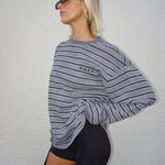 A woman wearing a gray and black horizontally striped long-sleeve sweatshirt with 'RAEF' printed on the chest, paired with black bike shorts, posing against a white textured wall.