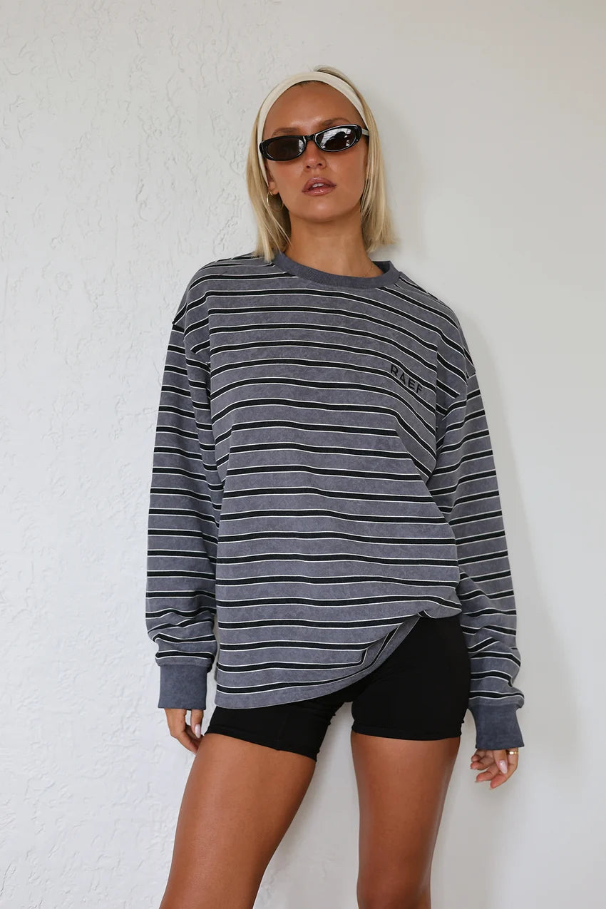 A woman modeling an oversized long-sleeve crewneck sweatshirt with thin horizontal gray and black stripes, paired with black shorts and sunglasses, standing against a white textured wall.