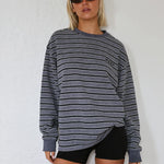 A woman modeling an oversized long-sleeve crewneck sweatshirt with thin horizontal gray and black stripes, paired with black shorts and sunglasses, standing against a white textured wall.