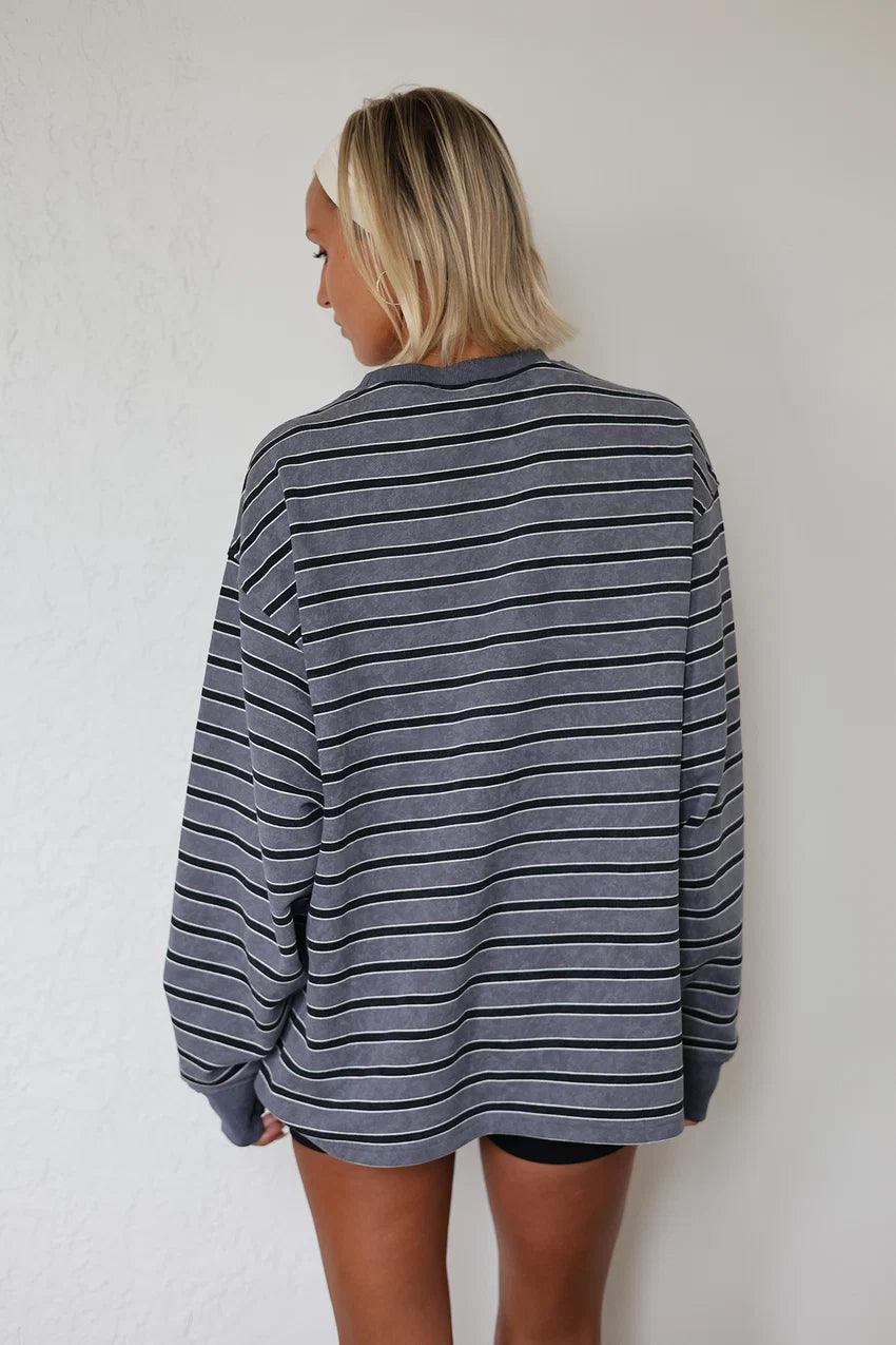 A woman with blonde hair is shown from behind wearing a gray and black horizontally striped long-sleeve shirt against a white textured wall.