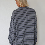 A woman with blonde hair is shown from behind wearing a gray and black horizontally striped long-sleeve shirt against a white textured wall.
