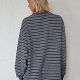 A woman with blonde hair is shown from behind wearing a gray and black horizontally striped long-sleeve shirt against a white textured wall.