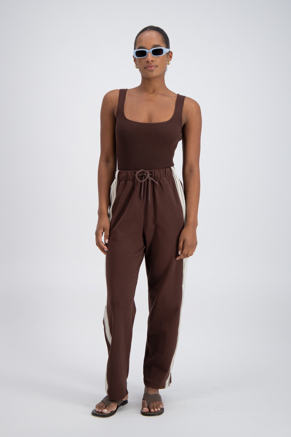 A model wearing chocolate brown trackpants with a relaxed fit, elastic waistband with drawstring, and pockets.