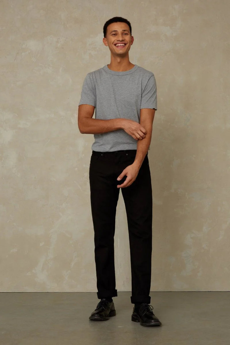 Man wearing a gray t-shirt and black pants standing against a beige wall.