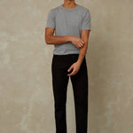 Man wearing a gray t-shirt and black pants standing against a beige wall.