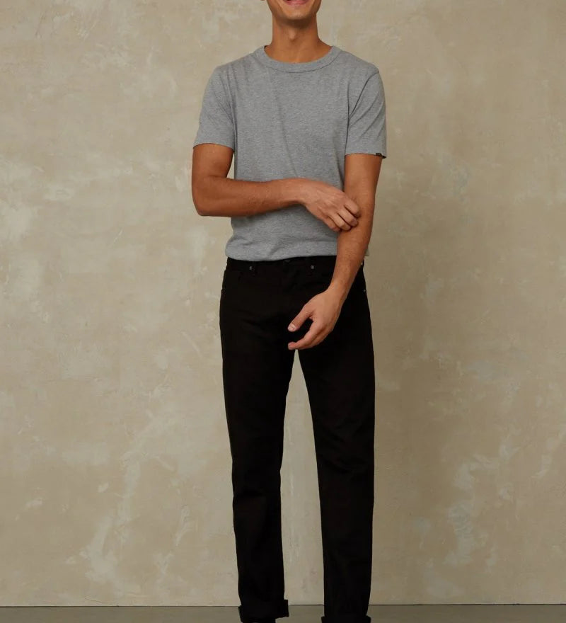 Man wearing a gray t-shirt and black pants standing against a beige wall.