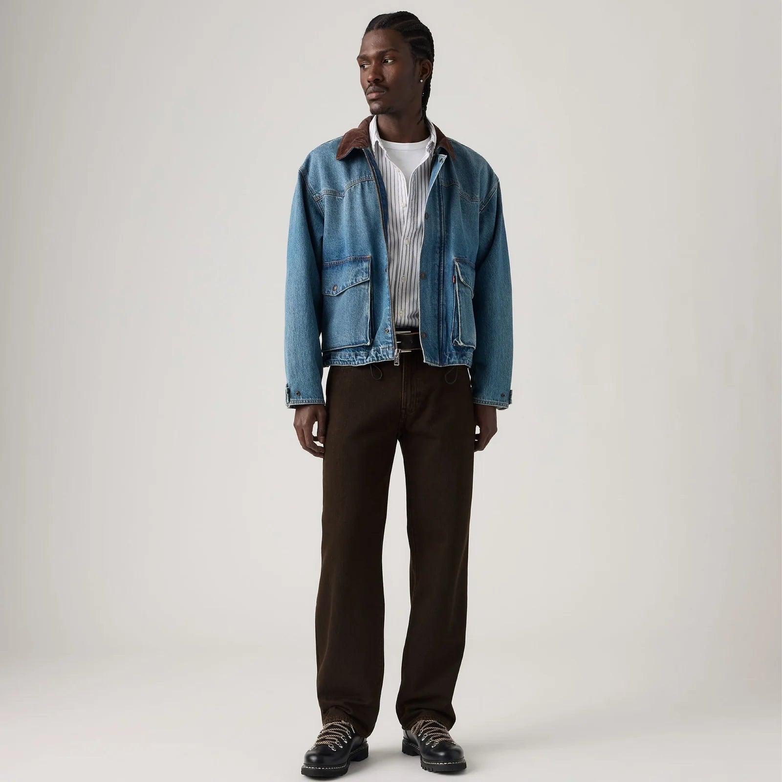A man modeling a light blue denim jacket with a brown suede collar, worn over a white striped shirt and dark brown trousers, standing against a neutral light gray studio background.