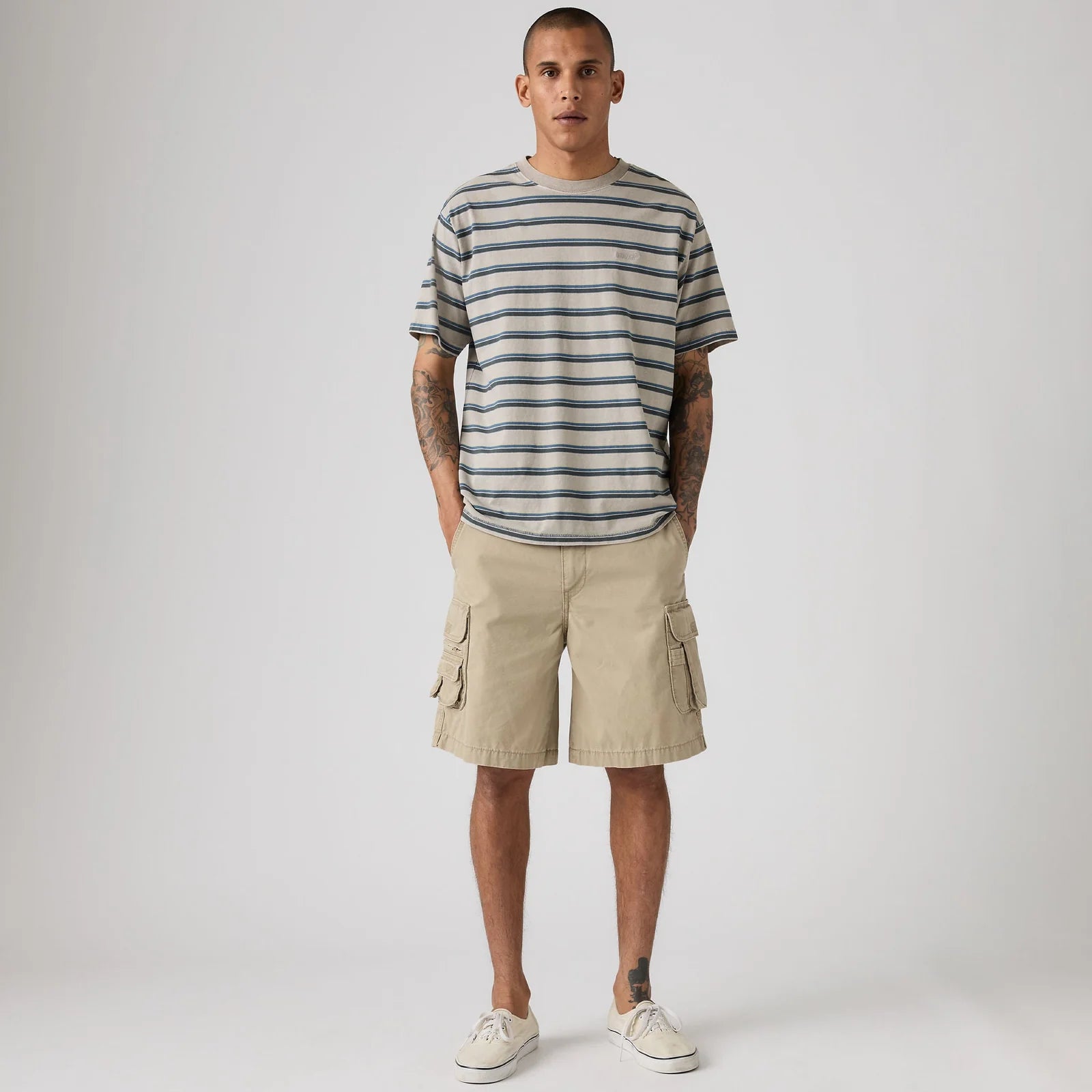 A man modeling a short-sleeve crewneck t-shirt with thin navy and beige horizontal stripes, paired with beige cargo shorts and off-white sneakers, against a light gray studio background.