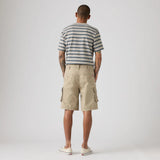 A man viewed from behind wearing a navy and white striped t-shirt, beige cargo shorts, and white sneakers, standing against a light gray studio background.