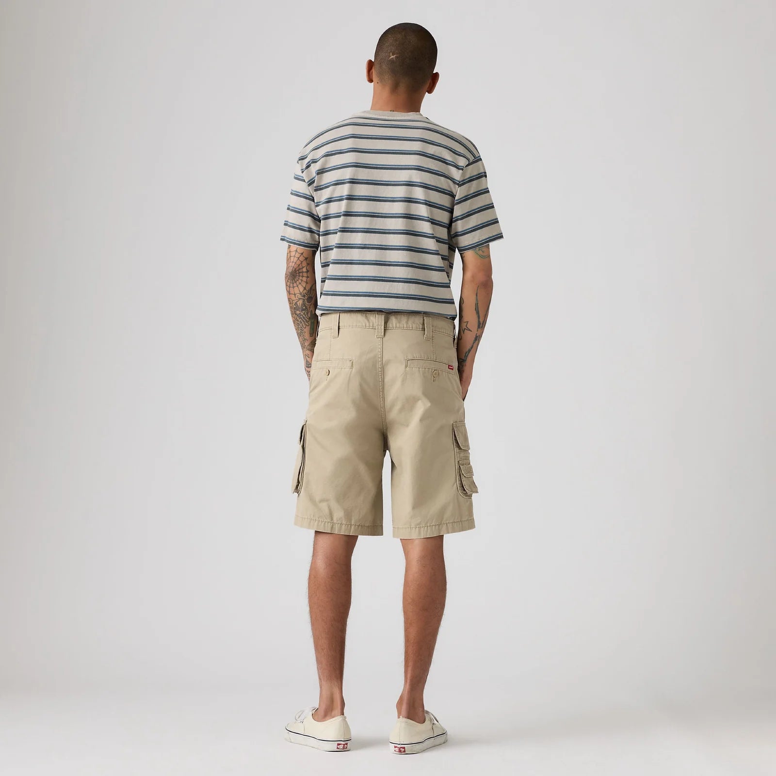 A man viewed from behind wearing a navy and white striped t-shirt, beige cargo shorts, and white sneakers, standing against a light gray studio background.