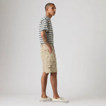 A man stands in profile wearing a short-sleeve striped t-shirt, beige cargo shorts, and white canvas sneakers against a plain light gray studio background.