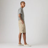 A man stands in profile wearing a short-sleeve striped t-shirt, beige cargo shorts, and white canvas sneakers against a plain light gray studio background.