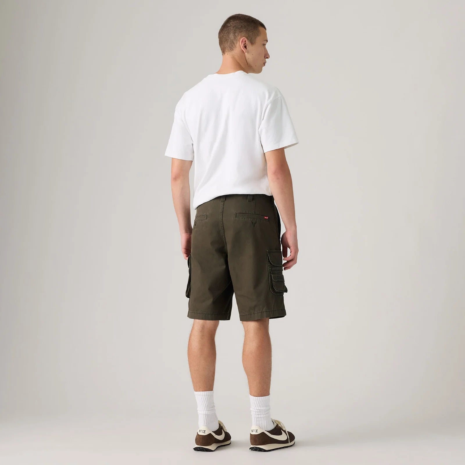 A man shown from the back wearing dark olive green cargo shorts, a white t-shirt, white socks, and brown sneakers, standing against a light gray studio background.