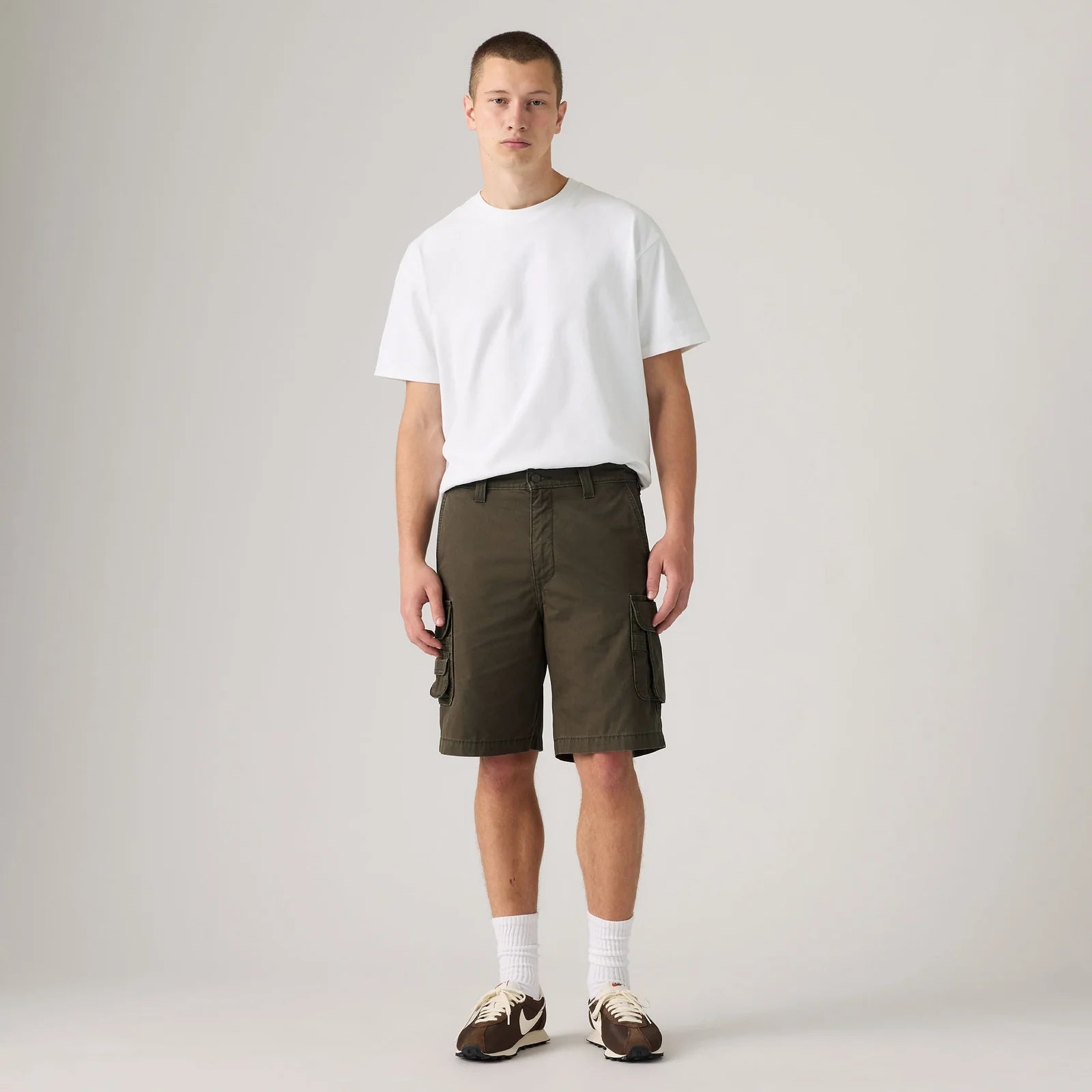 A male model stands against a light gray studio background wearing a plain white short-sleeve T-shirt, olive green cargo shorts, white socks, and brown sneakers.