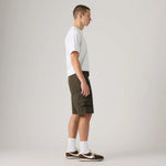 A man in profile wearing olive green cargo shorts, a white t-shirt, white socks, and brown sneakers, standing against a light gray studio background.