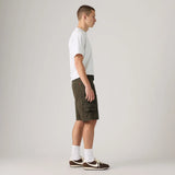 A man in profile wearing olive green cargo shorts, a white t-shirt, white socks, and brown sneakers, standing against a light gray studio background.