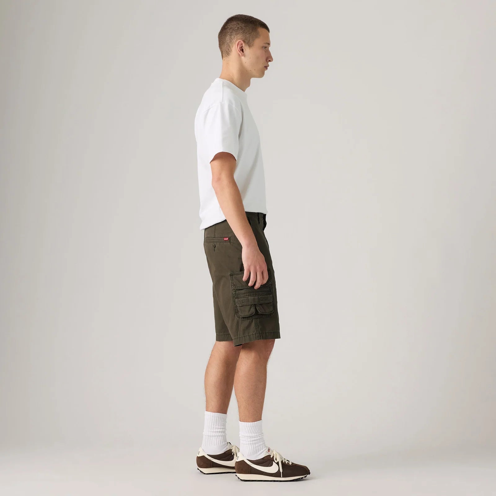 A man in profile wearing olive green cargo shorts, a white t-shirt, white socks, and brown sneakers, standing against a light gray studio background.