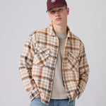 A young man wearing a brown and beige plaid flannel shirt over a beige t-shirt, blue jeans, and a maroon baseball cap, photographed against a solid light gray background.