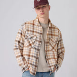 A young man wearing a brown and beige plaid flannel shirt over a beige t-shirt, blue jeans, and a maroon baseball cap, photographed against a solid light gray background.