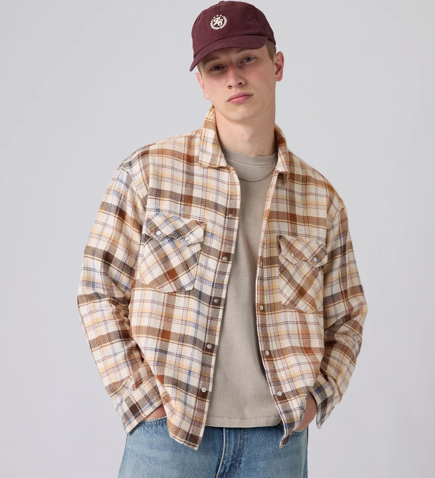 A young man wearing a brown and beige plaid flannel shirt over a beige t-shirt, blue jeans, and a maroon baseball cap, photographed against a solid light gray background.