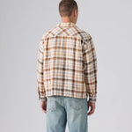 A man viewed from behind wearing a beige and brown plaid flannel shirt and light blue jeans, standing against a neutral gray studio background.