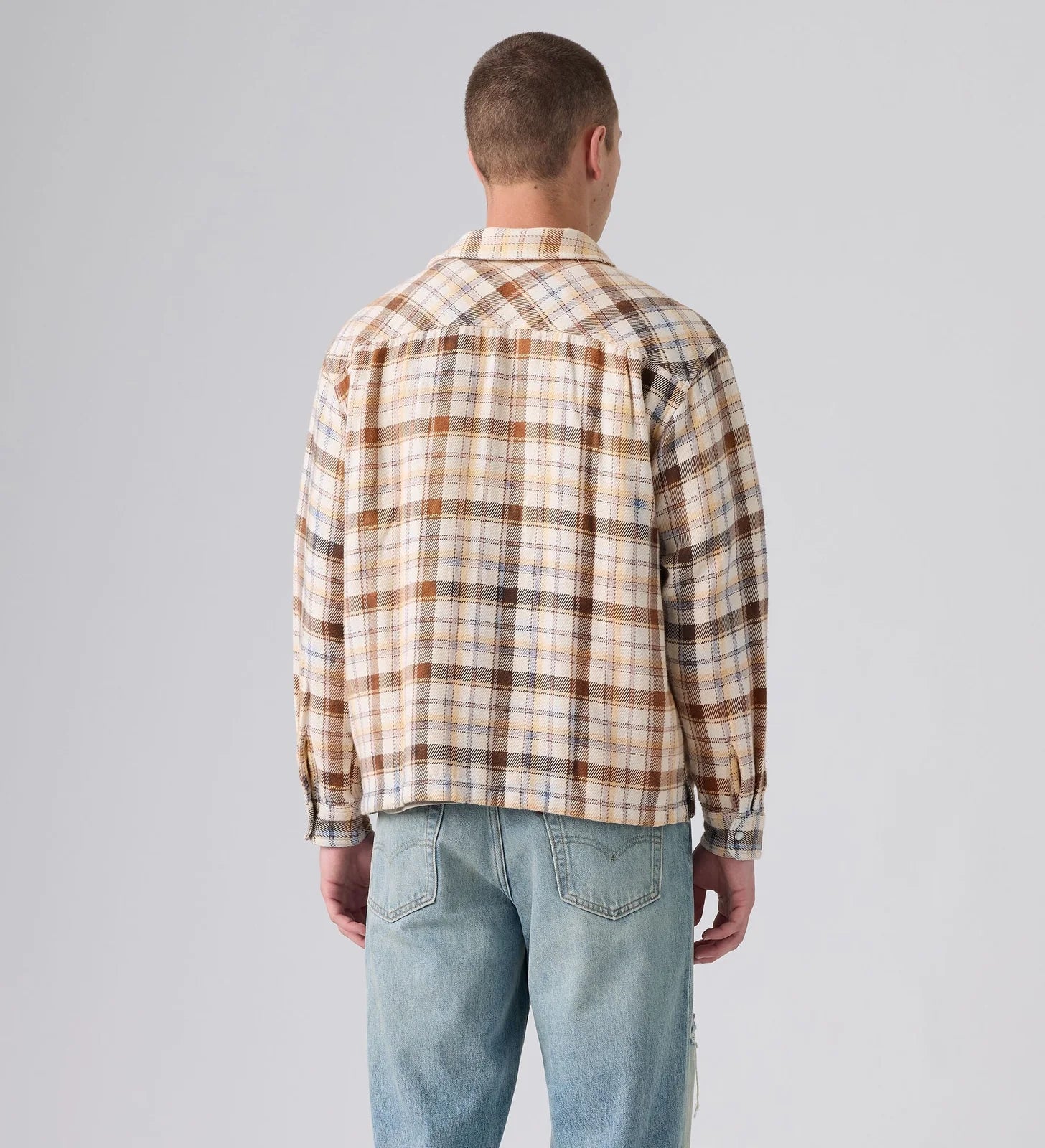 A man viewed from behind wearing a beige and brown plaid flannel shirt and light blue jeans, standing against a neutral gray studio background.