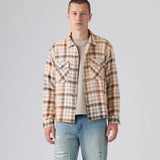 A male model wearing a beige and brown plaid overshirt over a light beige t-shirt and blue jeans, standing against a light gray studio background.