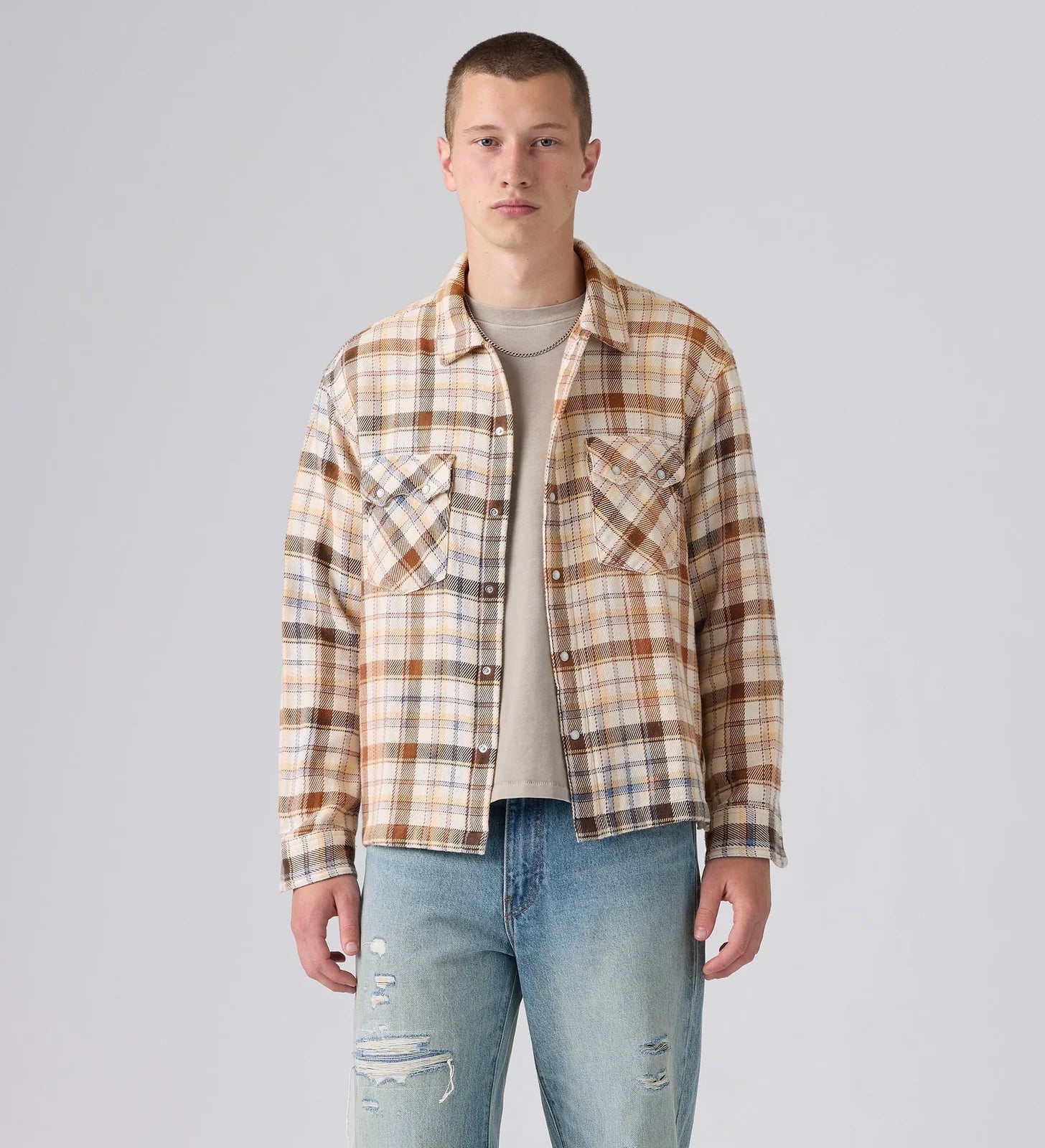 A male model wearing a beige and brown plaid overshirt over a light beige t-shirt and blue jeans, standing against a light gray studio background.