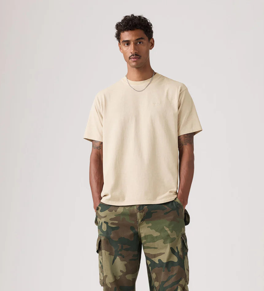 Man wearing a beige t-shirt and camouflage pants on a plain background