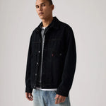 A man wearing a black denim trucker jacket over a black and white striped t-shirt and light blue jeans, standing against a light gray studio background.