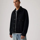 A man wearing a black denim trucker jacket over a black and white striped t-shirt and light blue jeans, standing against a light gray studio background.