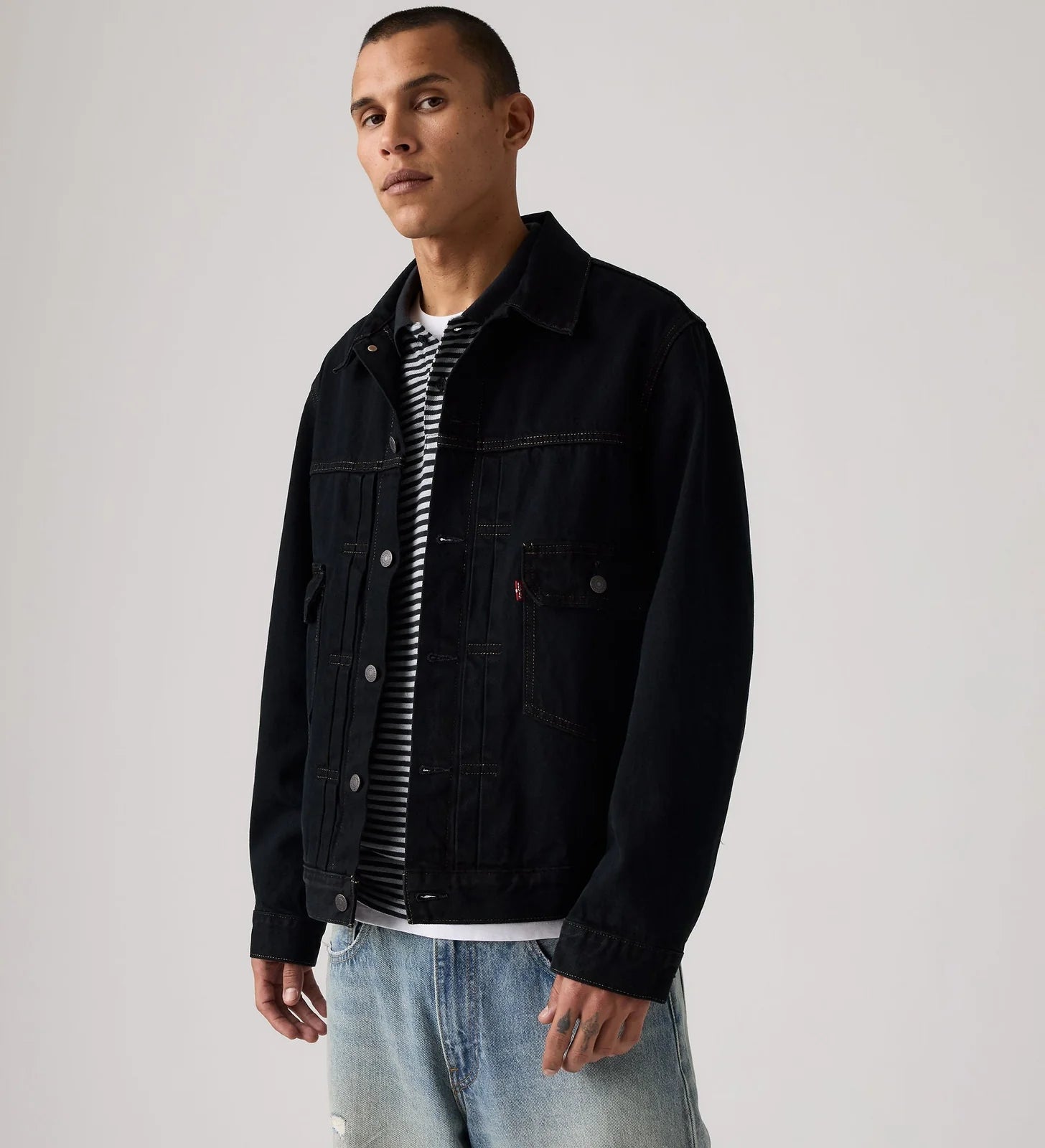 A man wearing a black denim trucker jacket over a black and white striped t-shirt and light blue jeans, standing against a light gray studio background.